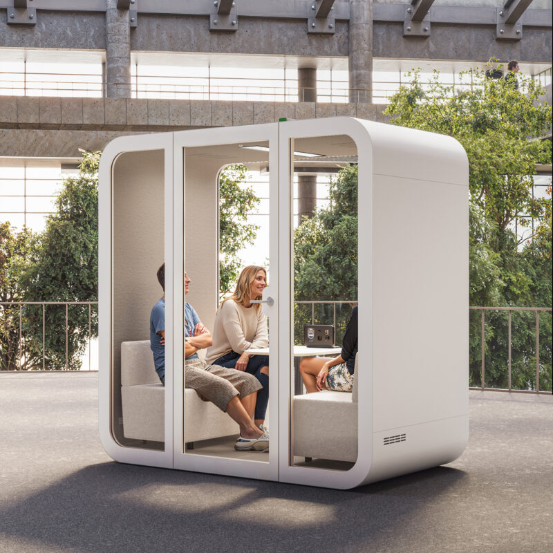 Three people relaxing inside a modern, soundproof white meeting pod with glass panels on an outdoor patio.